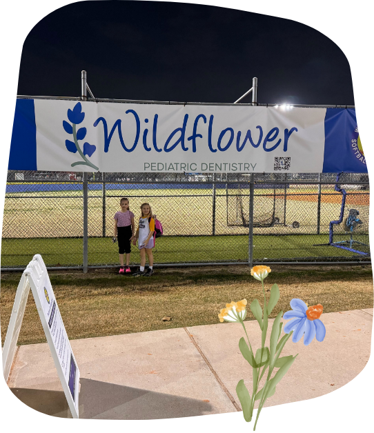 Two girls pose in front of a Wildflower sign, with vibrant flowers and lush plants in the background.  